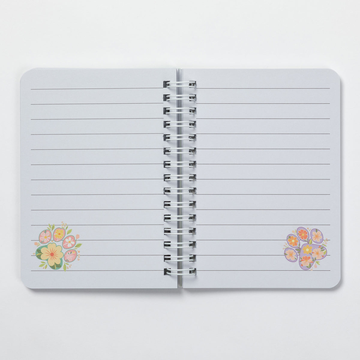 A lined notebook with a silver spiral binding, featuring small pastel floral illustrations in the bottom corners, shown open on a light gray background.