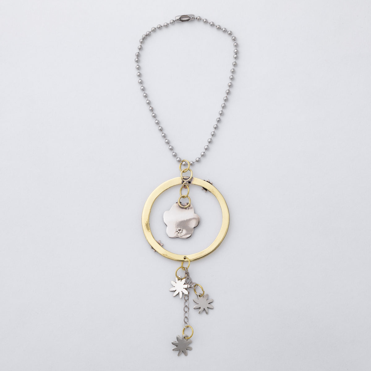 A silver-toned beaded necklace with a gold-toned circular frame and a central silver flower charm, featuring three dangling star and leaf charms, shown on a light gray background.
