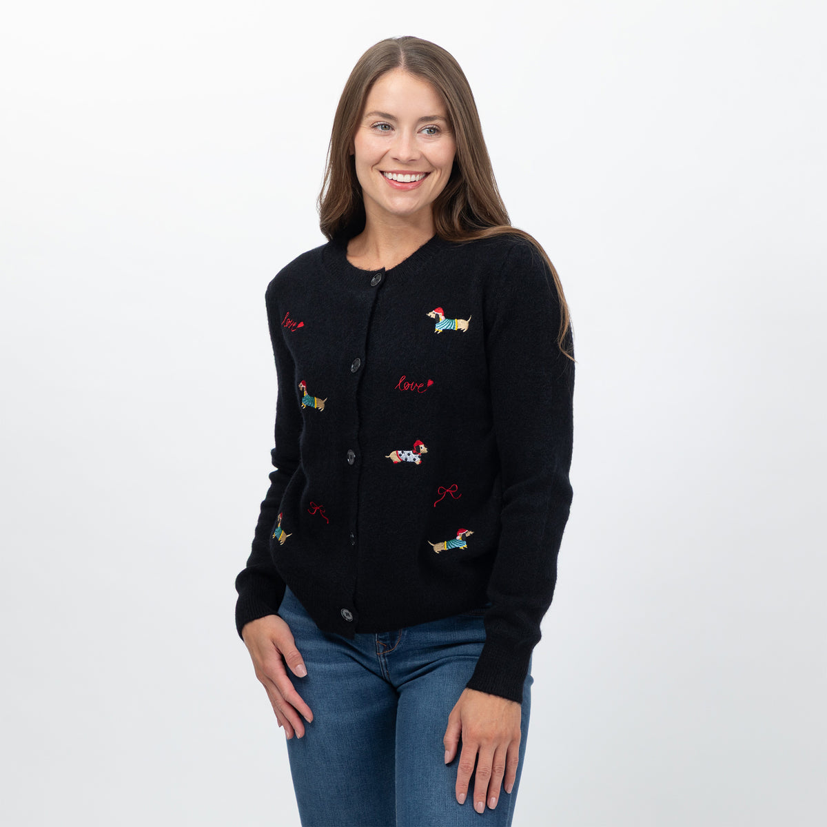 A woman wearing a black knit cardigan with colorful embroidered dog motifs and red script text, paired with blue jeans, photographed against a plain light gray studio background.