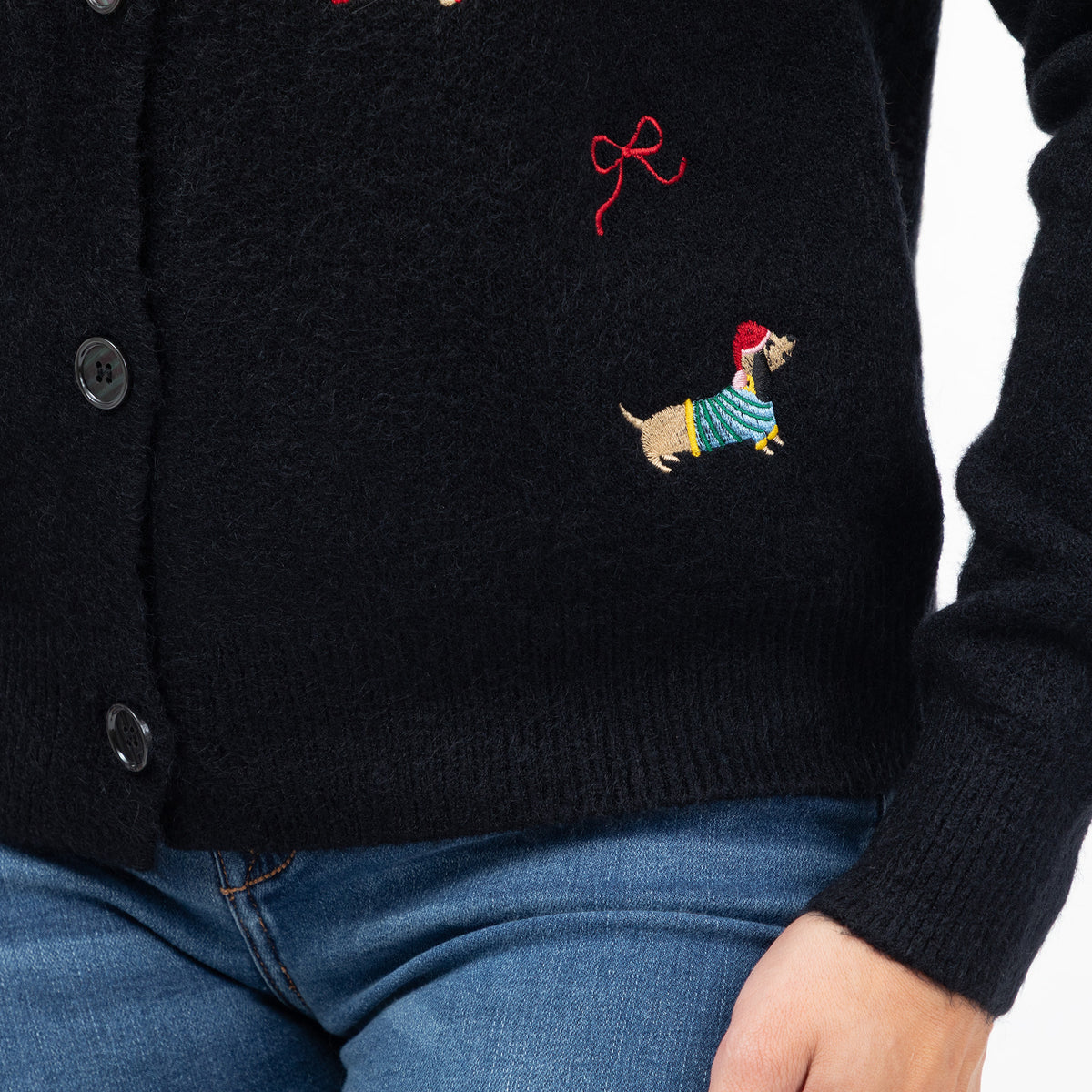 Close-up of a black knit cardigan sweater worn by a person, featuring embroidered details of a dachshund in a striped sweater and a red bow on the chest, paired with blue jeans.