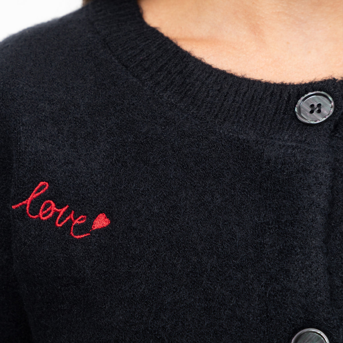 Close-up of a black knit cardigan sweater with red embroidered script reading 'love' and a small red heart on the chest, plus dark buttons.
