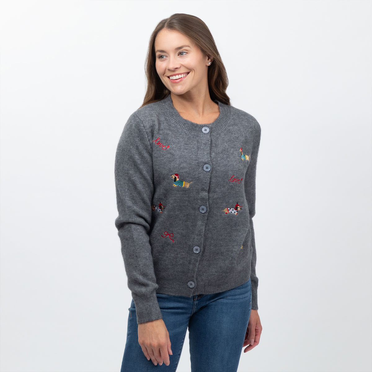 A woman wearing a gray button-front cardigan with colorful embroidered dog motifs, paired with blue jeans, photographed against a plain light gray studio background.