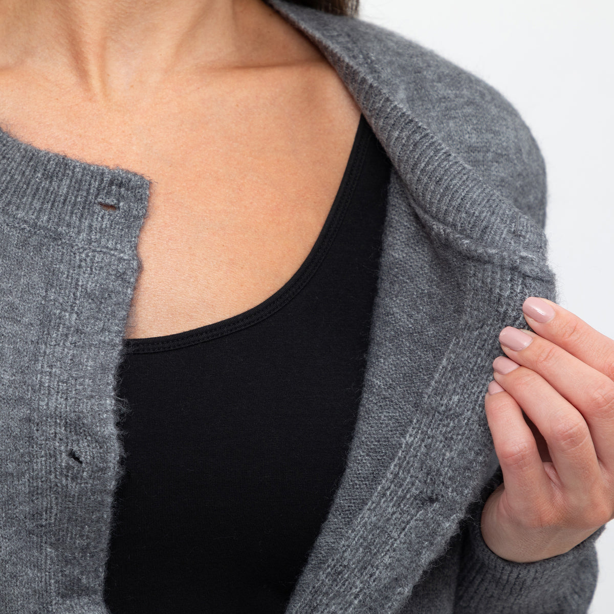 Close-up of a person wearing a heather gray knit cardigan over a black scoop-neck top, with one hand holding the cardigan open against a plain light background.