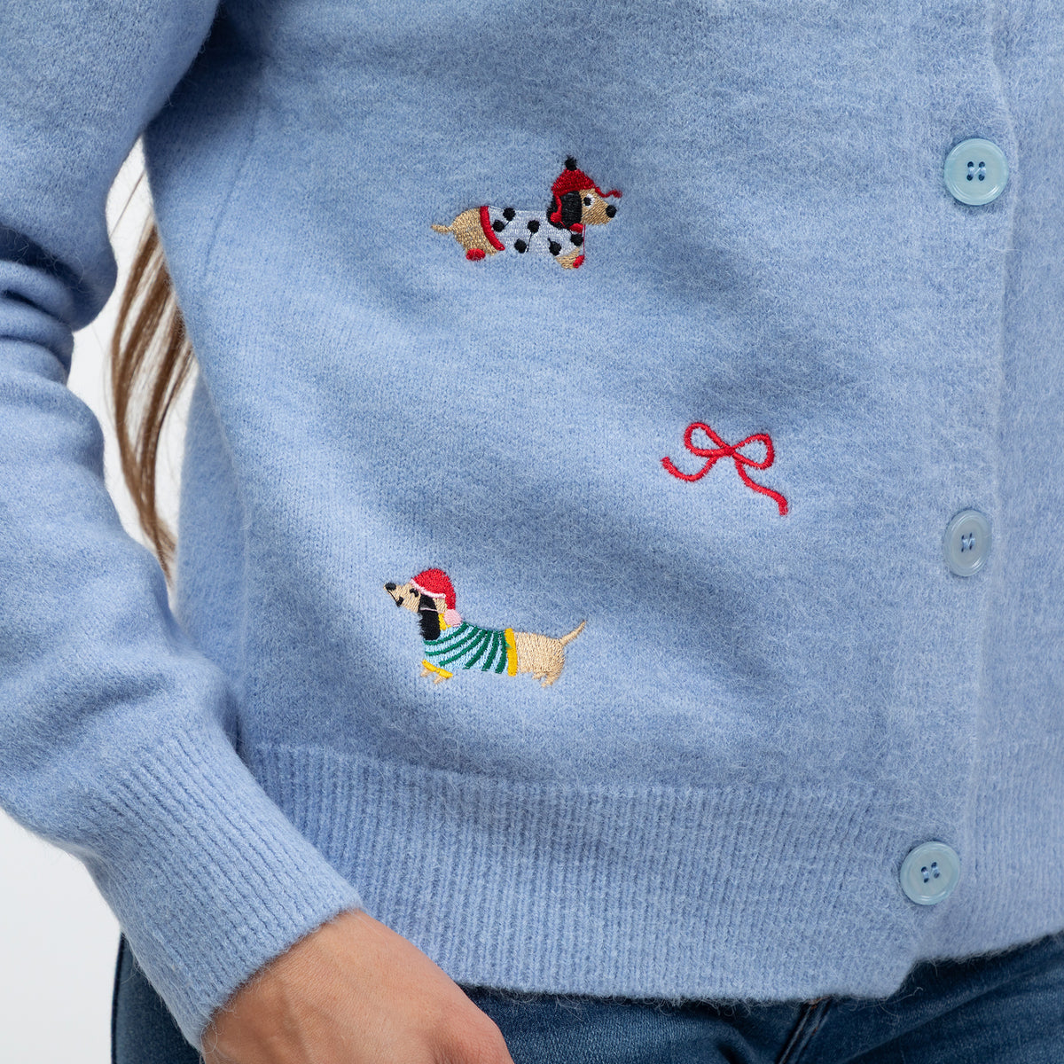 A close-up of a light blue knit cardigan worn by a person, featuring embroidered dachshunds in red and green outfits and a red bow motif on the chest.