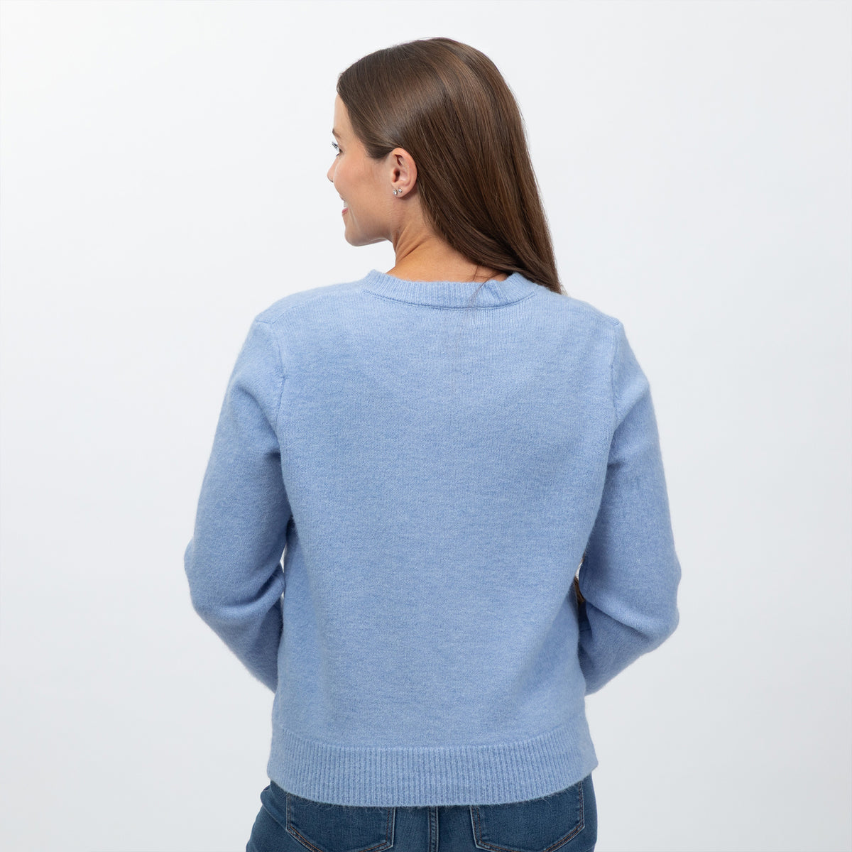 A woman viewed from the back wearing a light blue knit sweater and blue jeans against a plain white studio background.