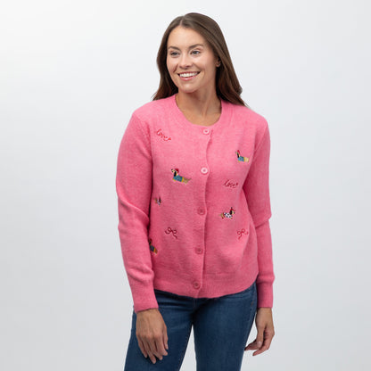 A woman wearing a pink button-up cardigan with embroidered dog motifs and the word 'love' on the front, paired with blue jeans, against a light gray studio background.