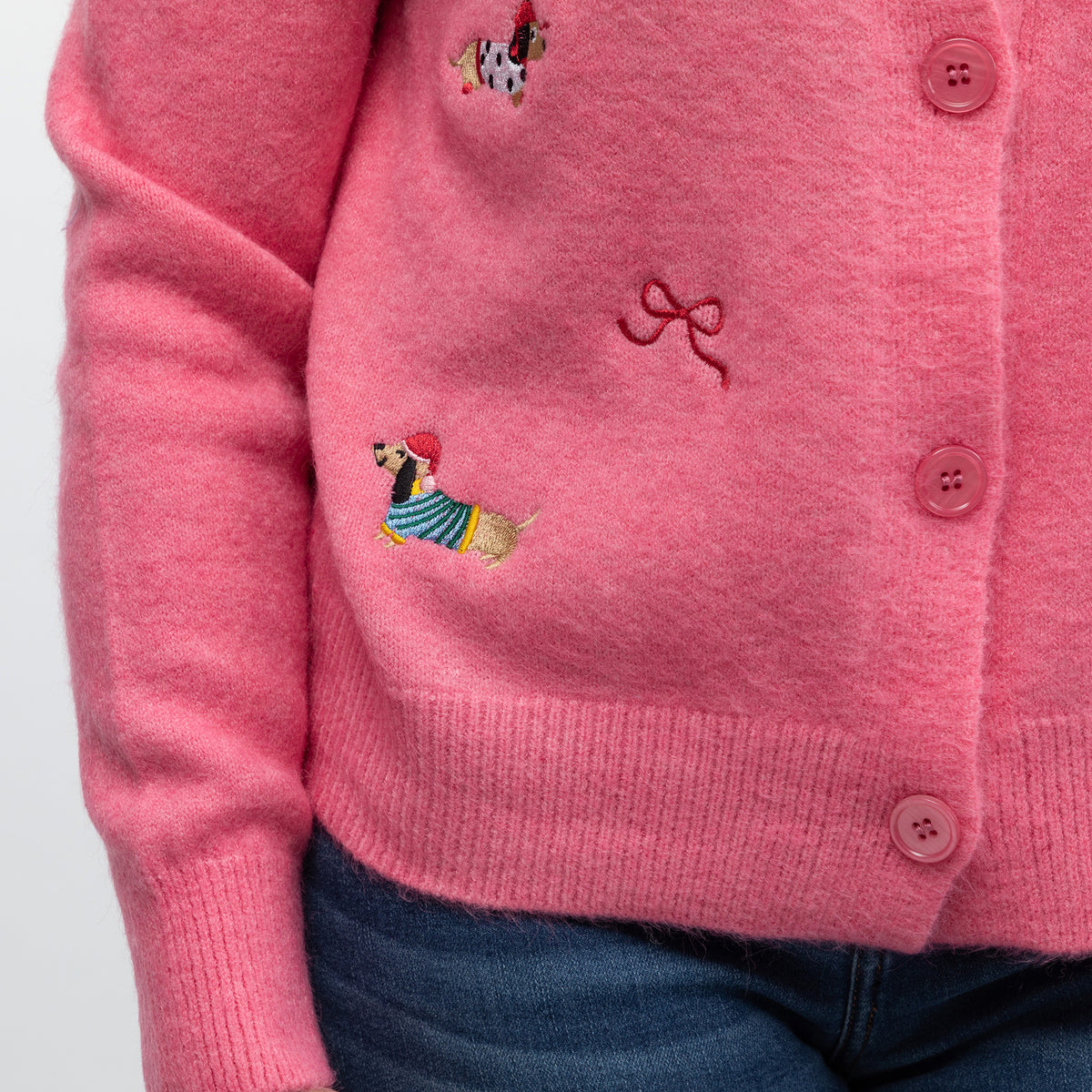 A close-up of a pink knit cardigan sweater worn by a person, featuring embroidered dog motifs and a bow detail on the chest, paired with blue jeans.