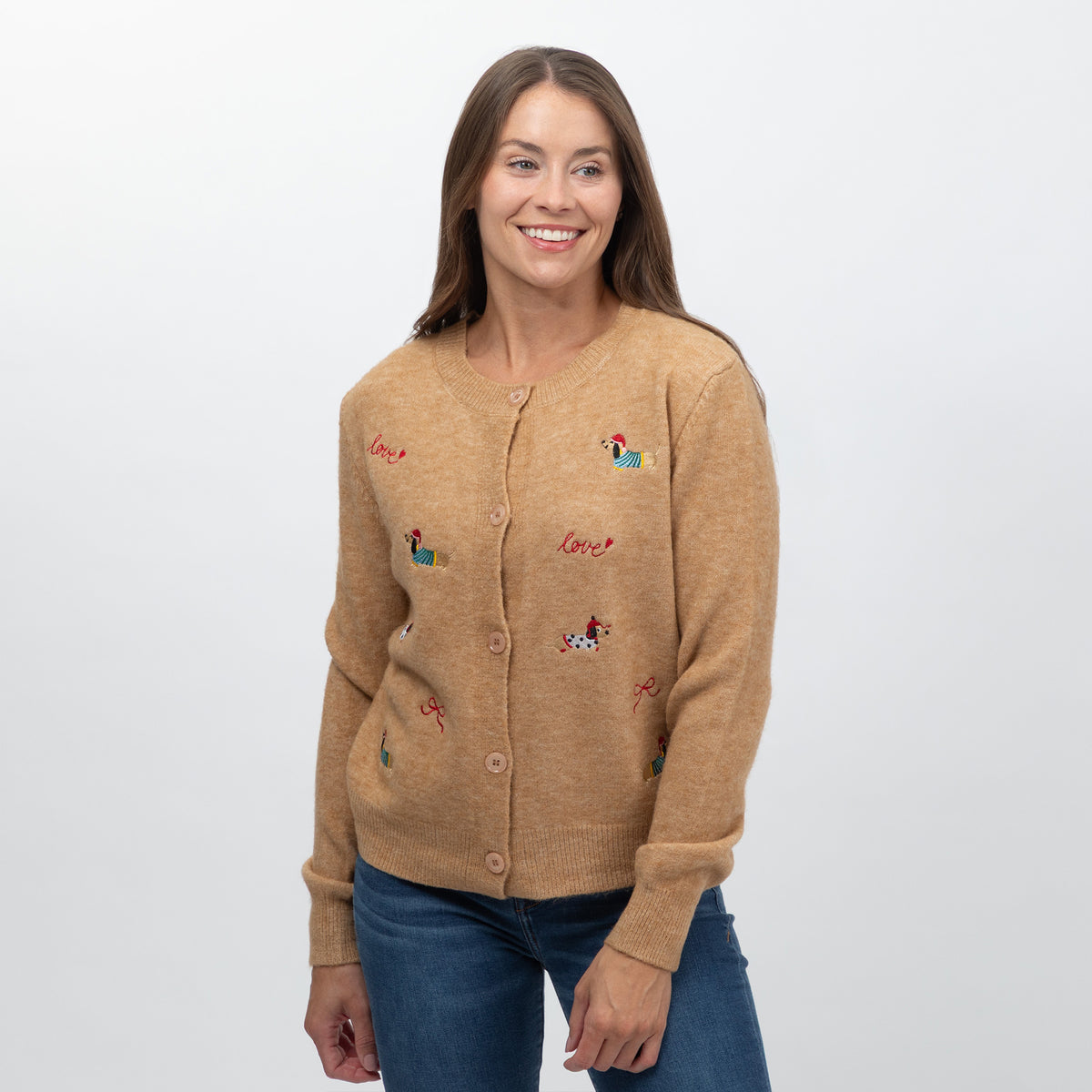 A smiling woman wearing a tan knit cardigan sweater with small embroidered dog and 'love' motifs, paired with blue jeans, against a plain light gray background.