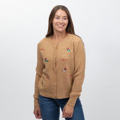 A smiling woman wearing a tan knit cardigan sweater with small embroidered dog and 'love' motifs, paired with blue jeans, against a plain light gray background.
