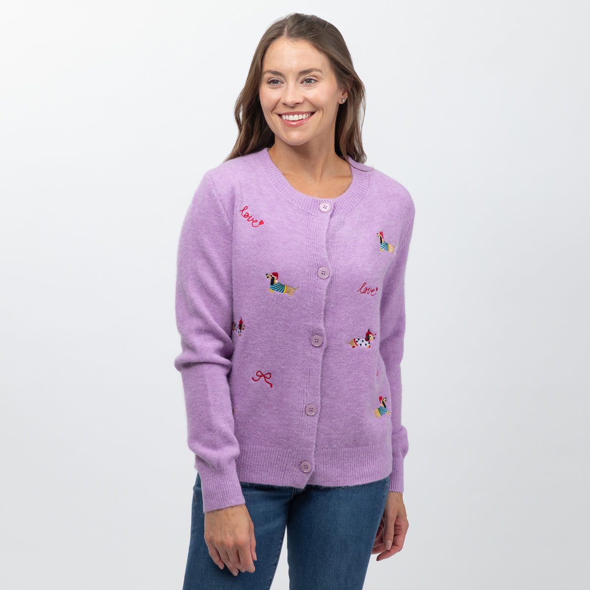 A woman wearing a light purple knit cardigan with colorful embroidered dog motifs and red script text, paired with blue jeans, against a plain light gray background.