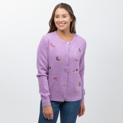 A woman wearing a light purple knit cardigan with colorful embroidered dog motifs and red script text, paired with blue jeans, against a plain light gray background.