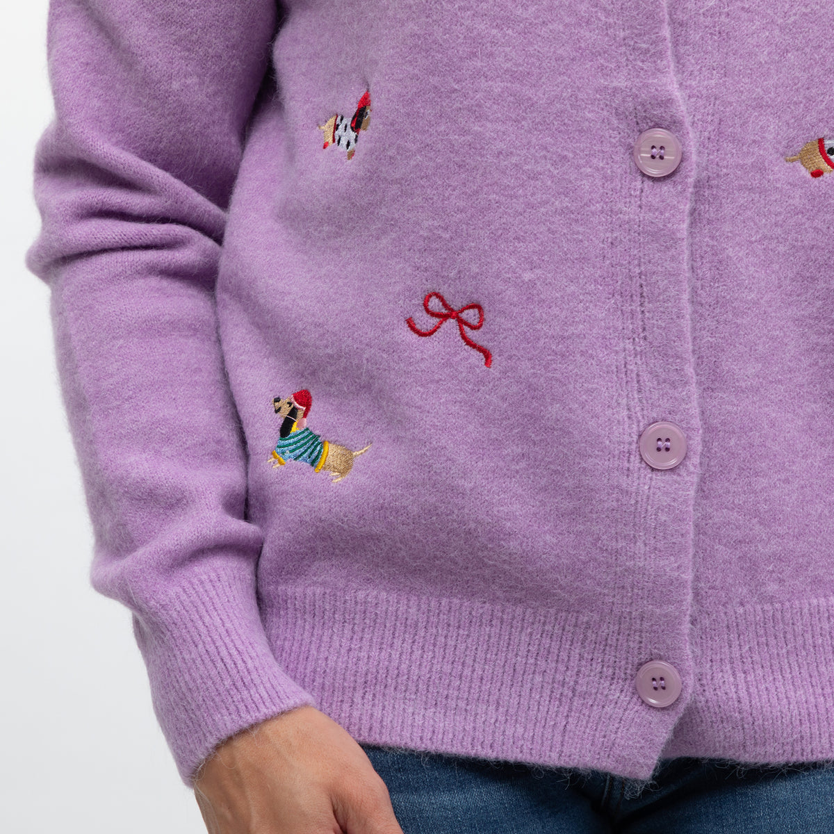 A close-up of a person wearing a lavender knit cardigan with embroidered dachshund dogs and a red bow motif, paired with blue jeans against a plain light background.