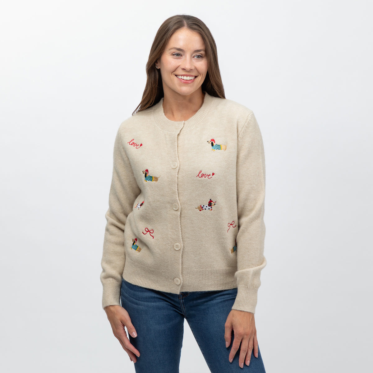 A woman wearing a beige knit cardigan with colorful embroidered dog illustrations and the word 'love' in red script, paired with blue jeans, against a plain light gray background.