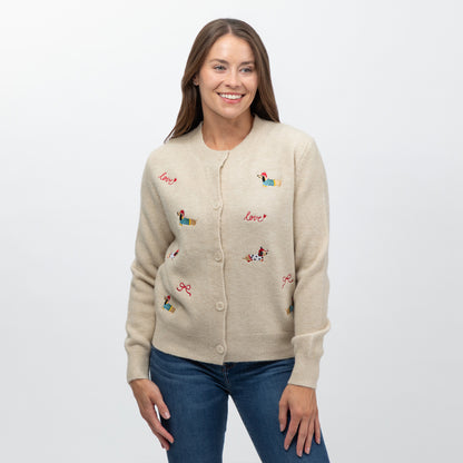 A woman wearing a beige knit cardigan with colorful embroidered dog illustrations and the word 'love' in red script, paired with blue jeans, against a plain light gray background.