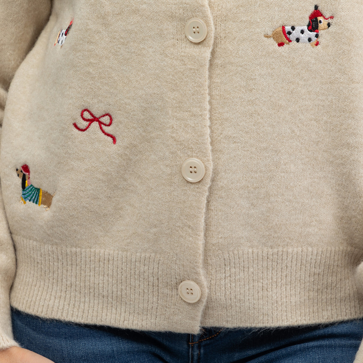 Close-up of a cream-colored knit cardigan sweater with embroidered dogs and a red bow motif, paired with blue jeans.