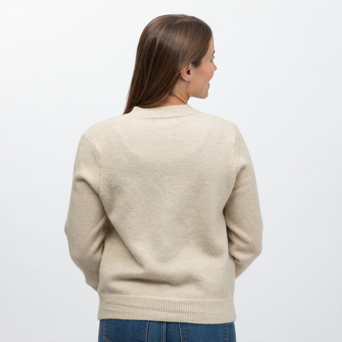 A woman viewed from the back wearing a light beige knit sweater and blue jeans against a plain white studio background.