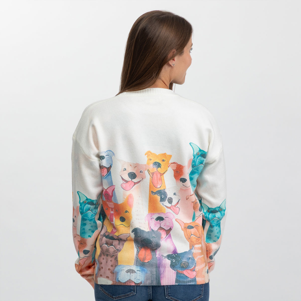 A woman viewed from behind wearing a cream-colored crewneck sweater with a colorful watercolor print of various dog faces across the back and sleeves, paired with blue jeans against a plain white background.