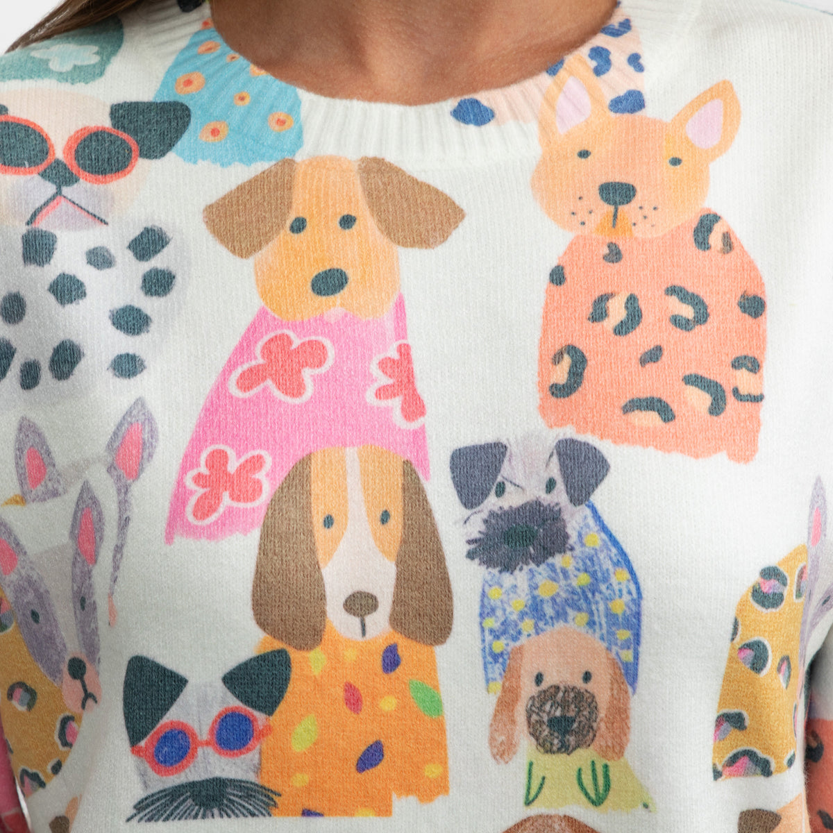 A close-up of a white knit sweater featuring a colorful cartoon dog print. The design includes various dog illustrations in pink, orange, yellow, blue, and black, with some dogs wearing patterned clothing.