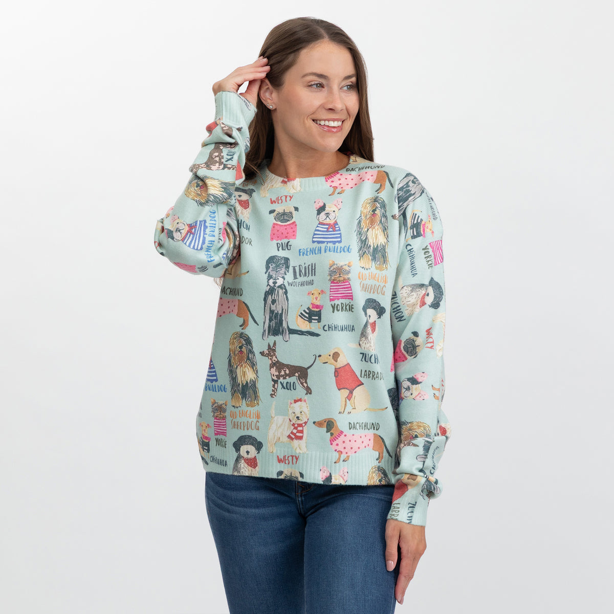 A woman modeling a light mint green long-sleeve crewneck sweater with a colorful all-over print of illustrated dogs and text labels, paired with blue jeans against a plain white studio background.