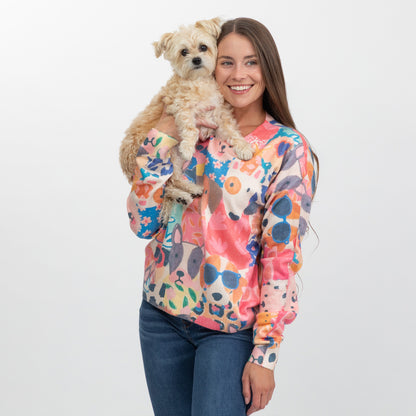 A woman holding a small fluffy dog, wearing a colorful long-sleeve crewneck sweater with a vibrant abstract floral pattern in pink, orange, blue, and yellow on a cream base, paired with blue jeans, against a plain light gray background.