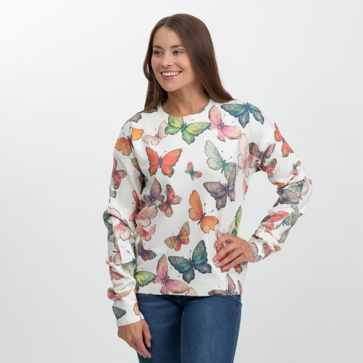 A woman modeling a cream-colored crewneck sweatshirt with a colorful butterfly print, paired with blue jeans, against a plain light gray background.