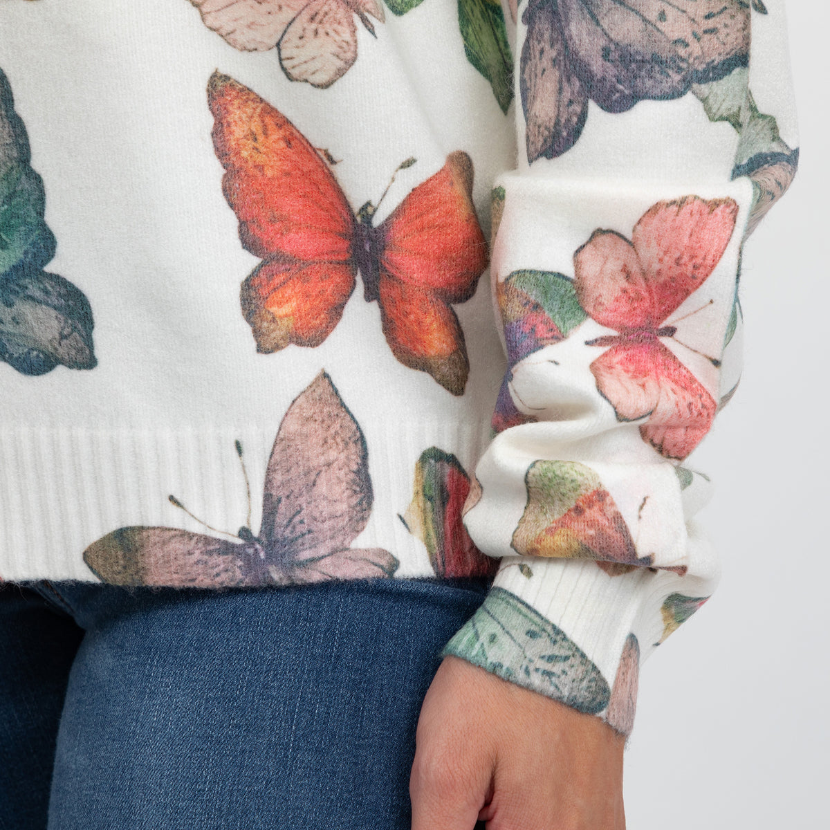 A close-up of a person wearing a white knit sweater with a large multicolor butterfly print and blue jeans, showing the sleeve cuff and hand.