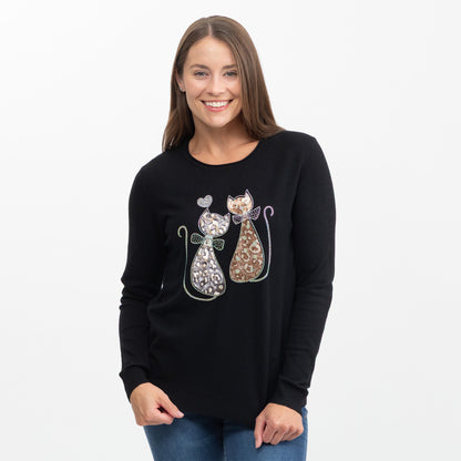 A woman wearing a black long-sleeve crewneck sweater with two cat figures made from rhinestones on the front, paired with blue jeans, against a plain white background.
