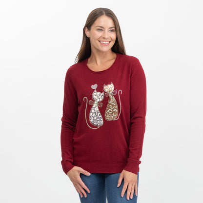 A woman wearing a maroon crewneck sweater with a graphic of two patterned cats, standing against a plain white background.