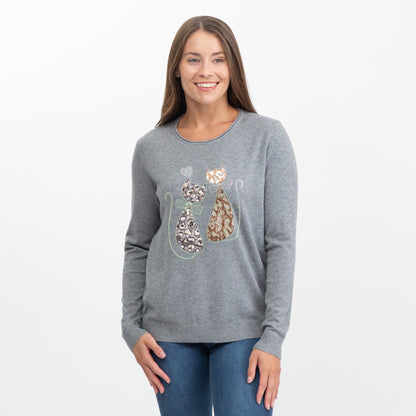 A woman modeling a heather gray long-sleeve crewneck sweater with a graphic of two cats on the front, paired with blue jeans against a plain white studio background.
