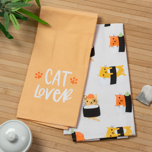 Two kitchen towels laid flat on a bamboo surface: one peach towel with white 'CAT LOVER' text and orange paw prints, and one white towel with a repeating cartoon cat and sushi print. A tea bag, plant leaves, and part of a white teapot are visible as styled props.