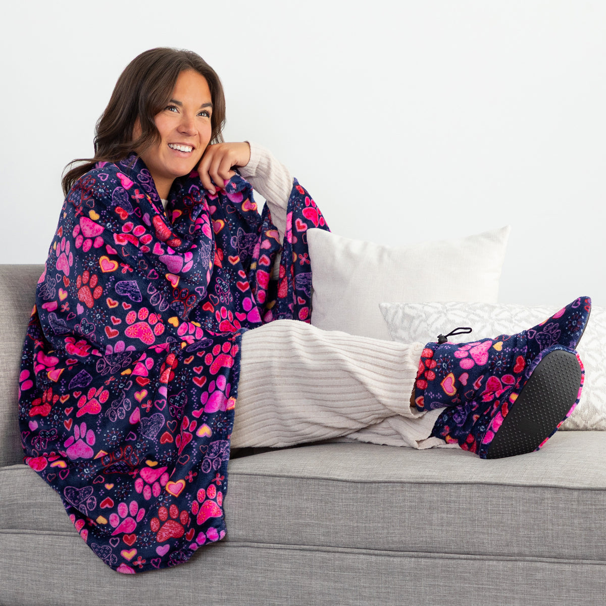 A smiling woman reclines on a gray sofa wrapped in a plush blanket and wearing matching slipper socks. The blanket and slippers feature a colorful pattern of pink hearts, purple paw prints, and orange accents on a dark navy background.