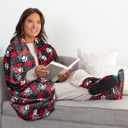 A woman sits on a gray couch reading a book, wrapped in a red and green plaid blanket with white paw prints, wearing matching plaid socks and light striped pajamas.