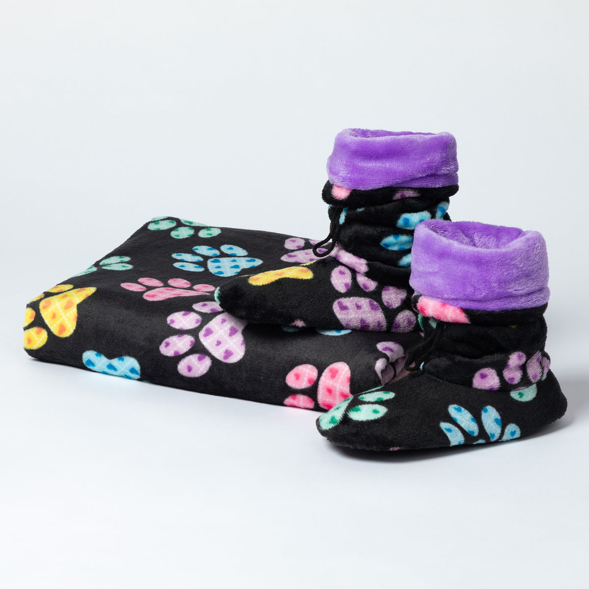 A pair of black bootie-style slippers with purple plush cuffs and a colorful paw print pattern, shown next to a matching folded black blanket on a light gray background.