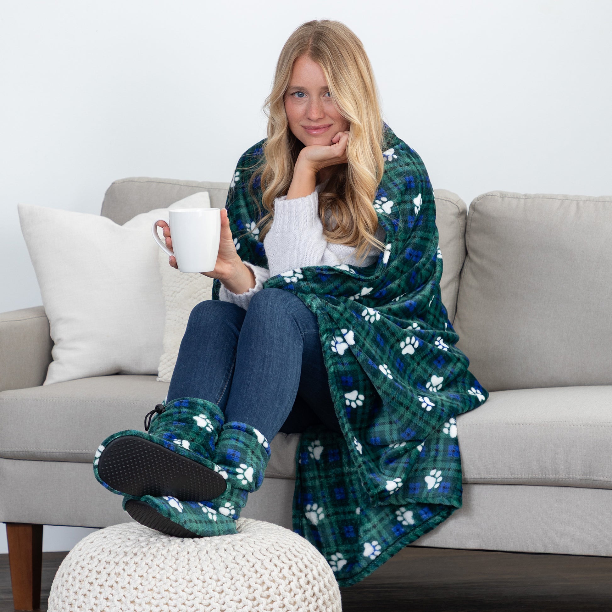 A woman sits on a light gray sofa wrapped in a green plaid blanket with white paw prints, holding a white mug. She is wearing matching green plaid slippers with black soles.