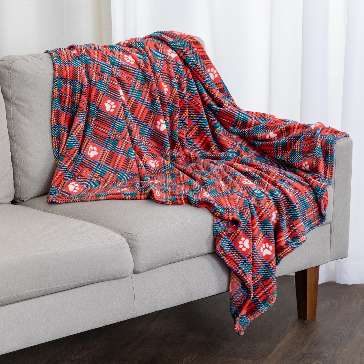 A red and teal plaid throw blanket with white paw print patterns draped over a light gray sofa in a living room setting.