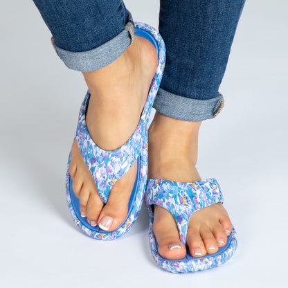 A pair of blue and purple EVA flip flops with marble paw garden paw print design, worn on a person's feet, standing on a white background