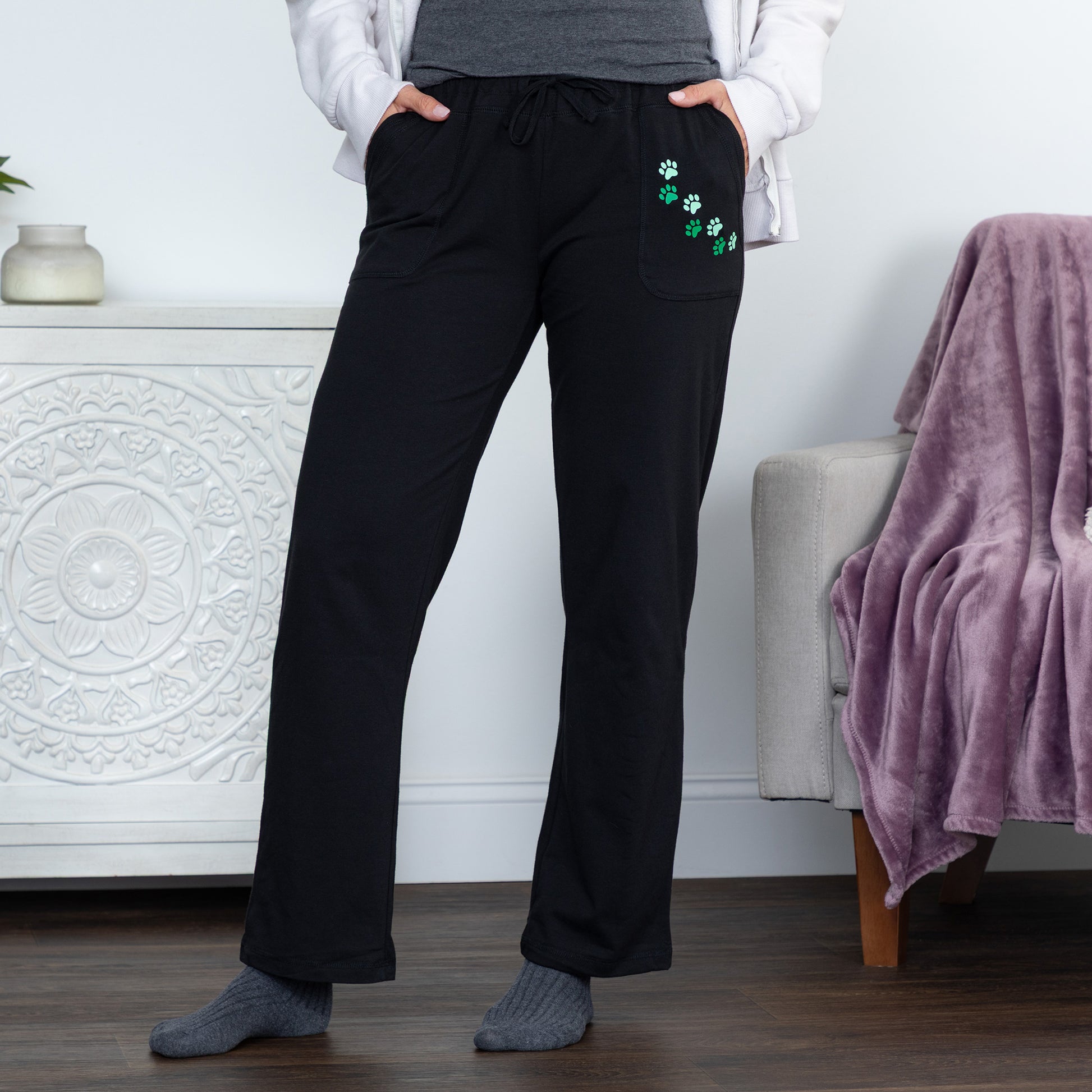 A person wearing black drawstring lounge pants with green shamrock embroidery on the pocket, standing indoors in a home setting with a white cabinet, wood floor, and a purple throw blanket on an armchair.