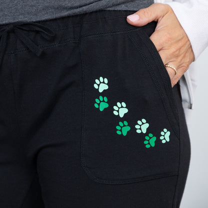 Close-up of a person wearing black drawstring sweatpants with a pocket decorated with a diagonal line of green paw prints, with a hand resting in the pocket.