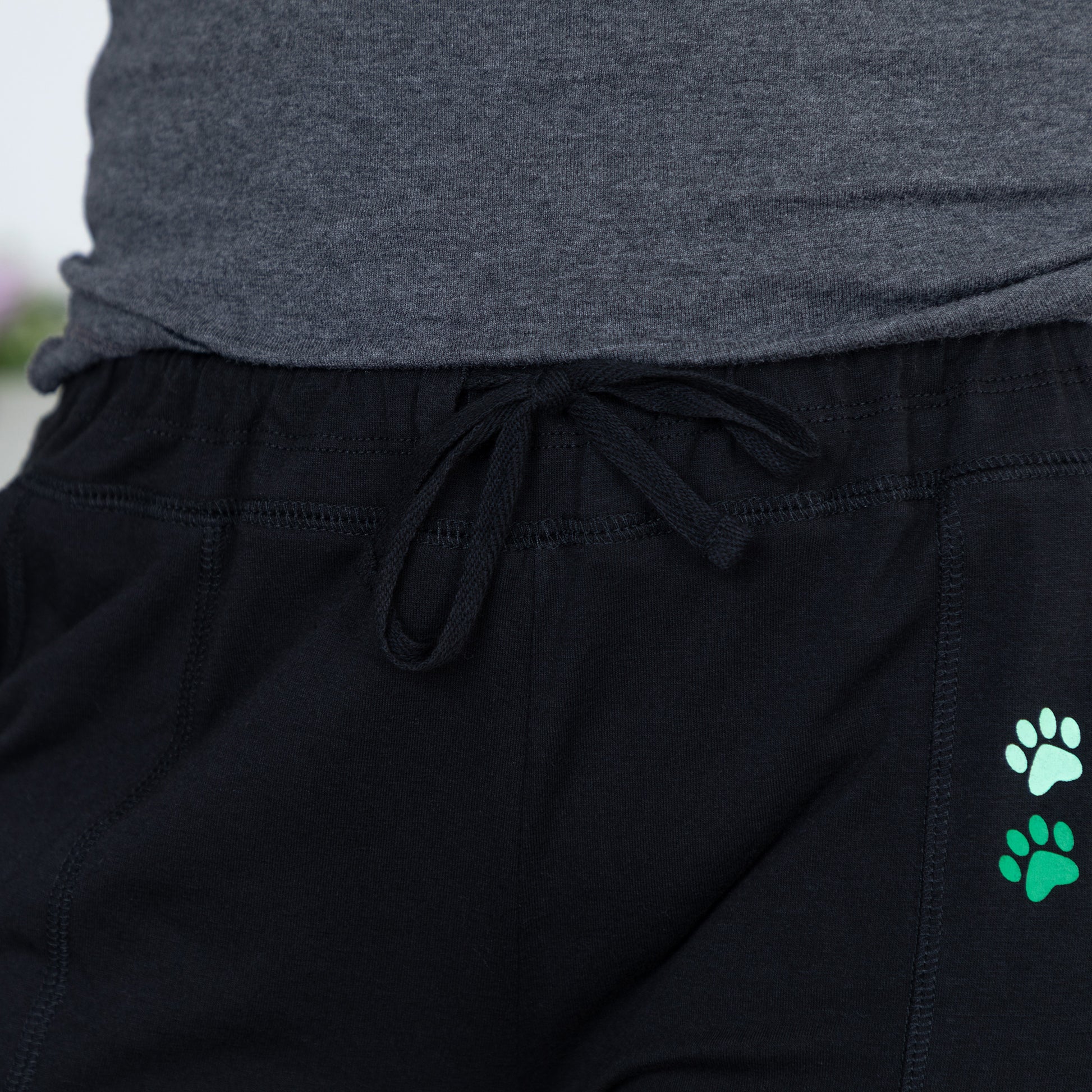 Close-up of black drawstring sweatpants with two small paw print graphics in mint green and bright green on the left leg, paired with a heather gray top.