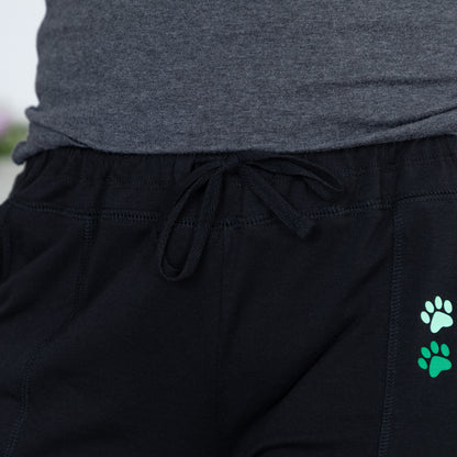Close-up of black drawstring sweatpants with two small paw print graphics in mint green and bright green on the left leg, paired with a heather gray top.