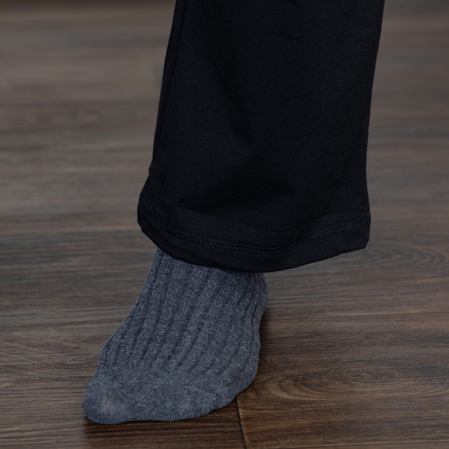 A close-up of a person's foot wearing a ribbed gray sock with black pants, standing on a dark wood floor.