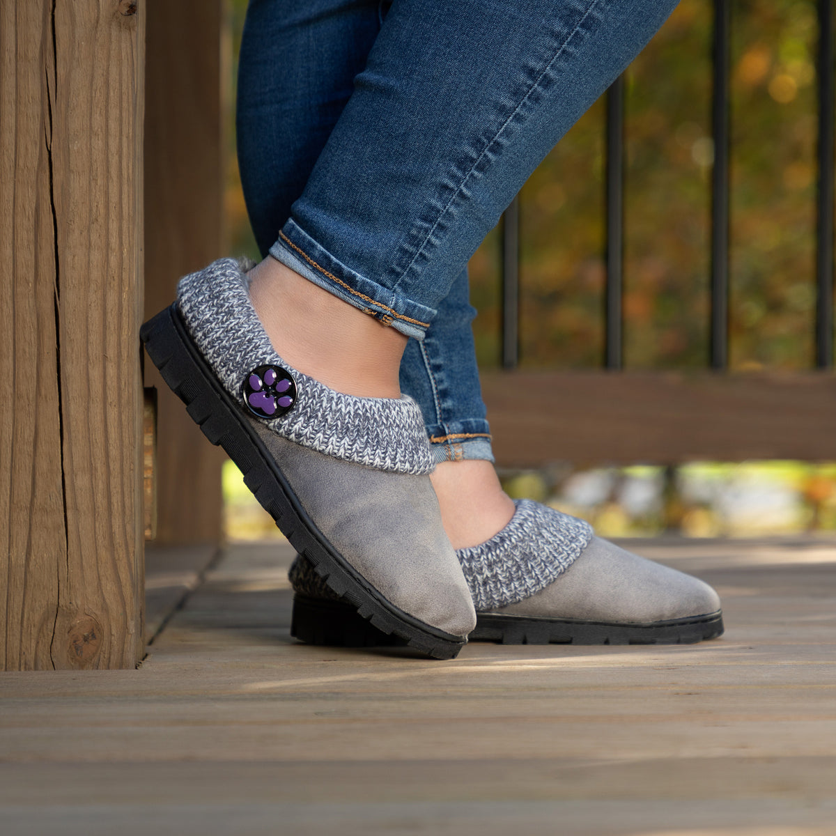 A person wearing gray slip-on clogs with gray knit trim and a purple decorative accent, standing on a wooden deck.