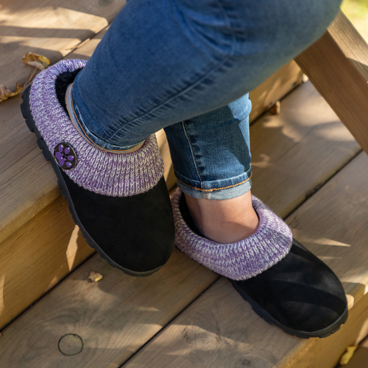 A person wearing black slip-on slippers with a purple knit trim and a decorative purple flower button, shown sitting on wooden outdoor steps.