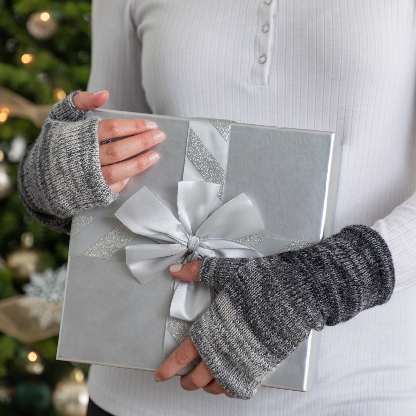 Person holding a silver gift box with a bow, wearing gray fingerless gloves, against a blurred Christmas tree background.