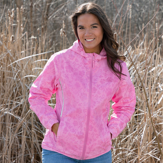 Smiling woman in a pink jacket stands among tall grasses outdoors.