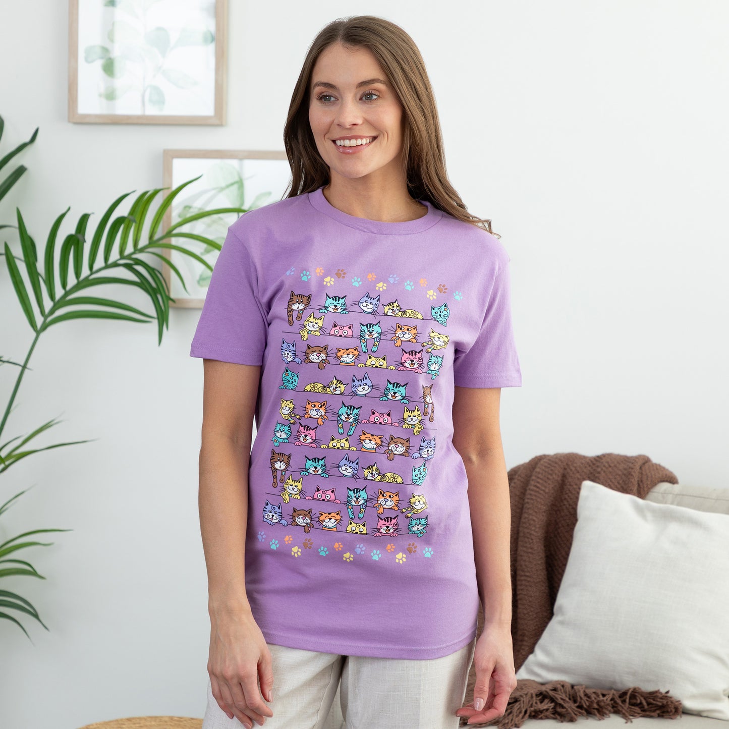 Woman wearing a purple t-shirt with colorful cat pattern in a room with a plant and couch.