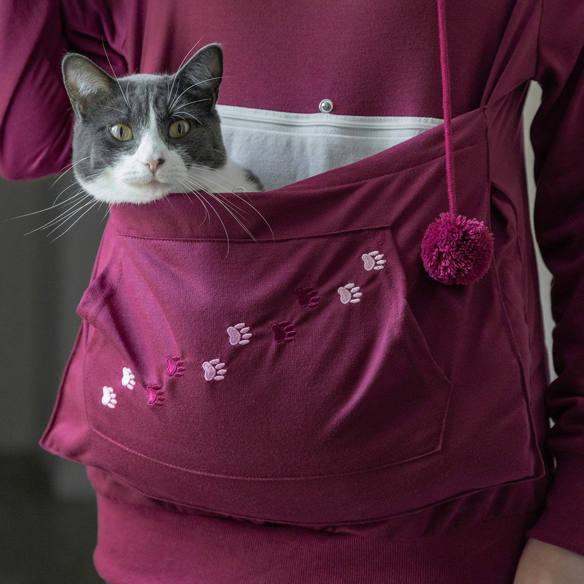 A gray and white cat is peeking out from a burgundy pet pouch worn on a person's sweatshirt. The pouch has embroidered paw prints and a matching pom-pom drawstring, shown against a matching burgundy hoodie.