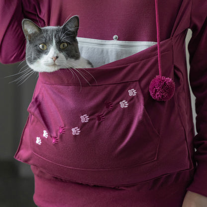 A gray and white cat is peeking out from a burgundy pet pouch worn on a person's sweatshirt. The pouch has embroidered paw prints and a matching pom-pom drawstring, shown against a matching burgundy hoodie.