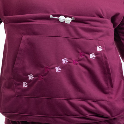 Close-up of a burgundy fleece jacket featuring a front kangaroo pocket with embroidered paw prints in shades of pink, purple, and white, secured with a silver metal snap button closure.