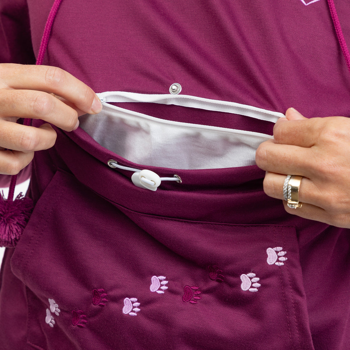 Close-up of a person's hands demonstrating the interior pocket and drawstring closure of a burgundy athletic hoodie with small pink paw print embroidery.